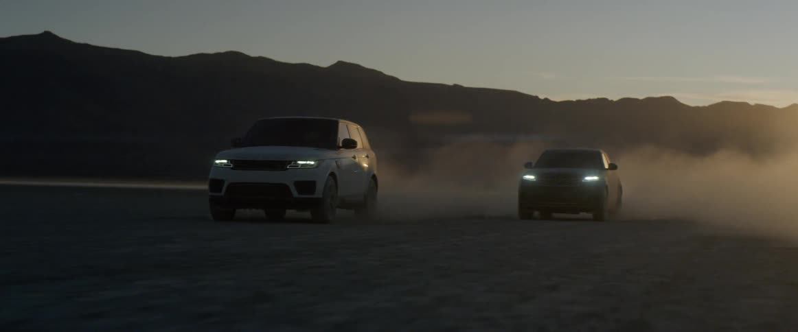 The vehicles race forward across the dirt plain with their headlights on
