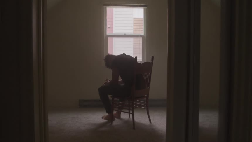 A man is sitting in a chair in an otherwise empty room looking downwards with his hands folded