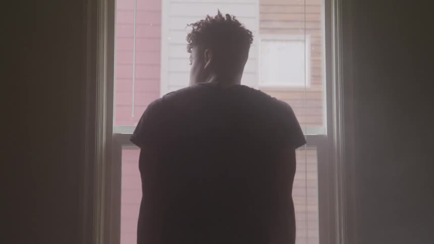 A young black teenager stands and looks out the window