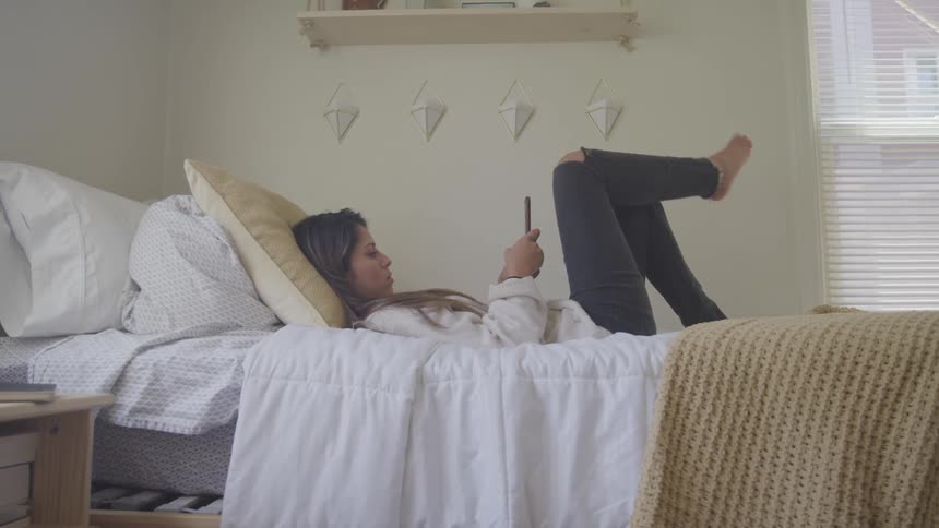 A teenage female lays in her bed while looking at her smart phone screen