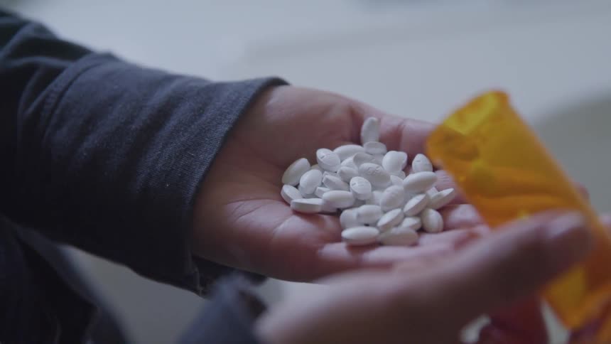 Woman pours a bottle of white oval pills into her hand leaving one left in the clear plastic orange bottle then closes her hand slightly over the medication