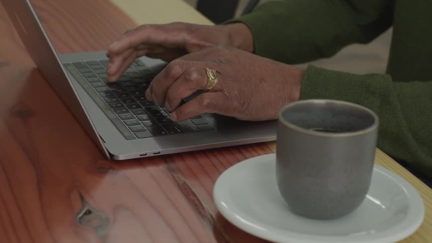 A man types on a laptop with a cup of coffee on the table beside him