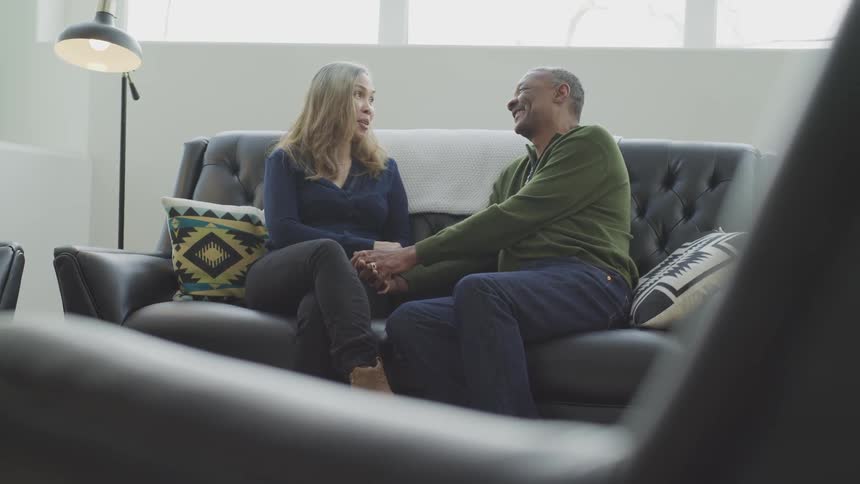 A man and woman smile while holding hands and talking on a couch