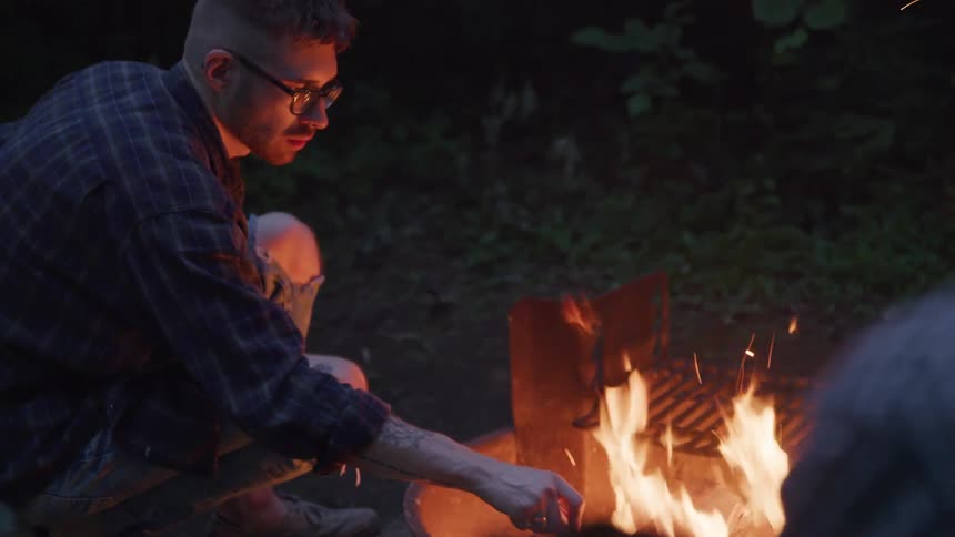 Male tends to a fire burning in an outdoor fire pit with a wooden stick as a female watches