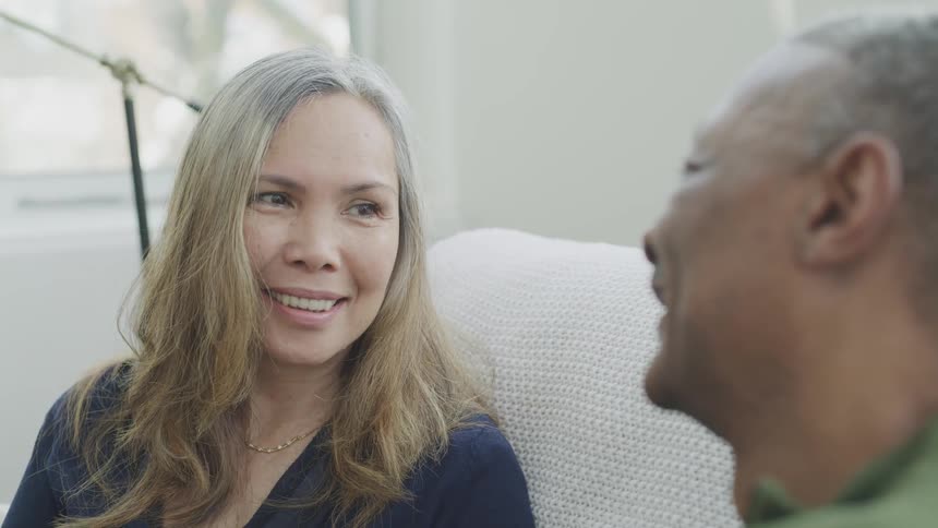 Black man and blonde woman sitting on the couch look at each other and smile in the living room