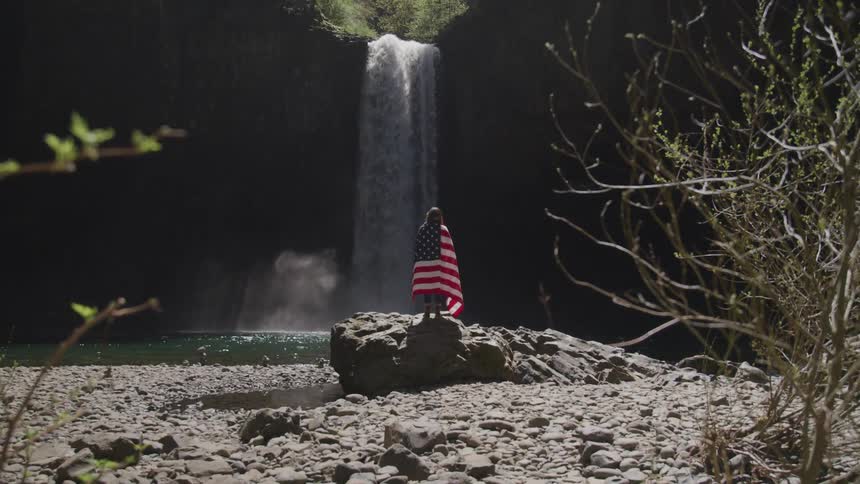 A person with an american flag draped around their shoulders standing on a large rock before a waterfall