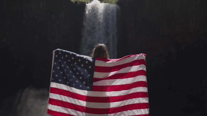 A woman holds an american flag behind her standing by a waterfall