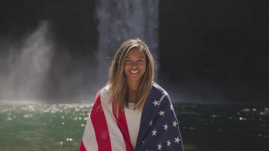 Woman stands draped in american flag in front of waterfall and pool