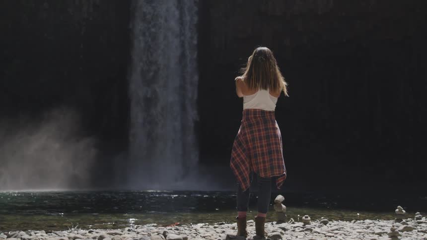 A woman with a plaid shirt tied around her waist takes a picture of a waterfall
