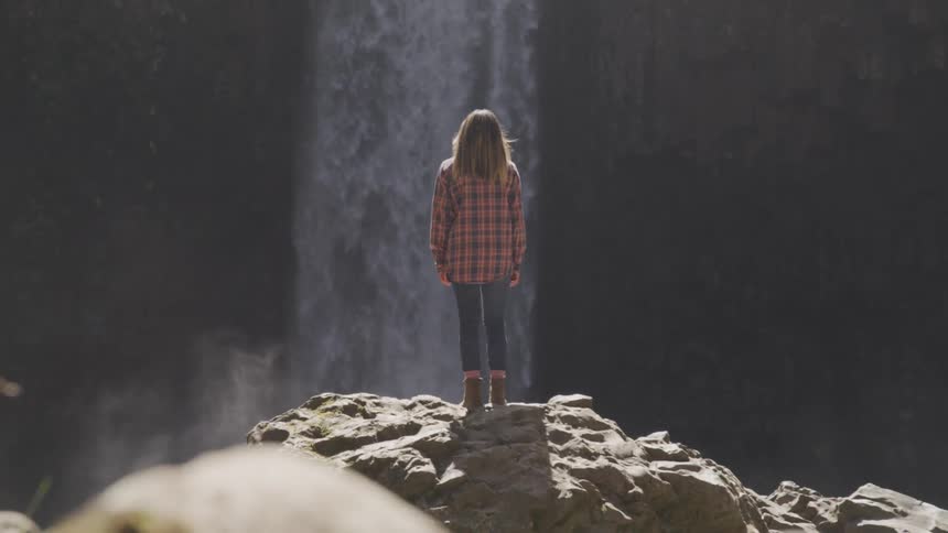 A woman stands on a rock looking at a flowing waterfall
