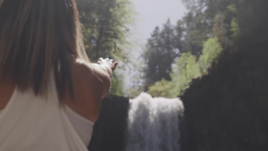 A woman slowly raising a hand towards a waterfall in the wilderness