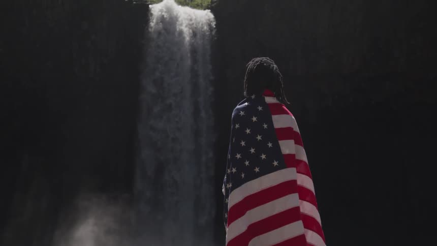 A person stands at the bottom of a waterfall with an american flag around their shoulders