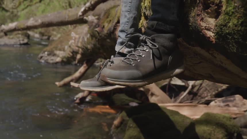 A person sitting on a log that spans a creek lets their feet dangle below them