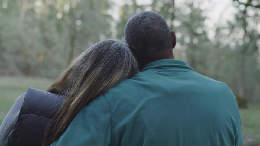 A man and woman cuddle outside near trees