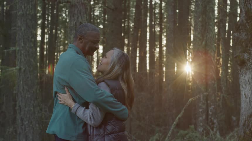 Black man and blonde woman hold each other while the sun streams through the trees in the forest