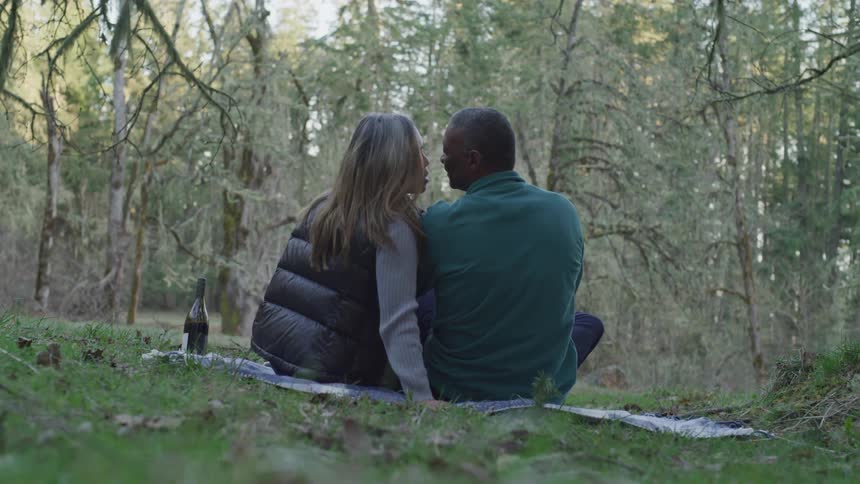 Black man and blonde woman sitting on the grass kiss each other in the woods