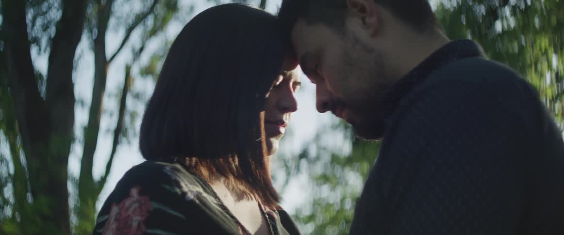 A man and woman stand face to face with foreheads together on a sunny day