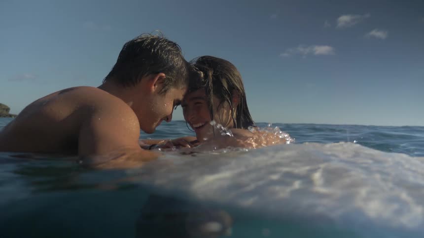 Man and woman float facing each other over surfboard