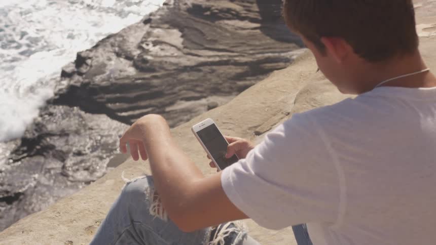 Young man with cellphone sitting in small cliff next to the ocean