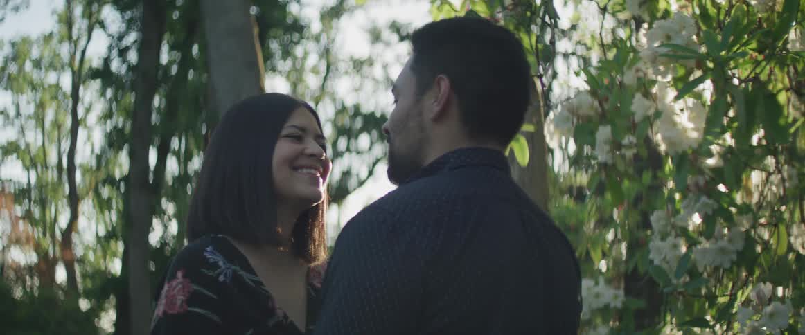 A couple stands face to face and smile near blossoming trees and shrubs