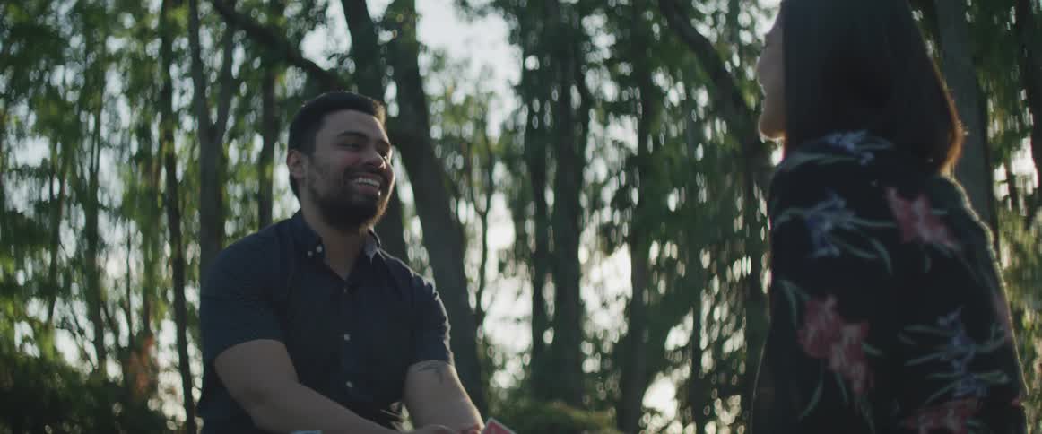 Man and woman lean toward each other laughing as sun shines through surrounding trees