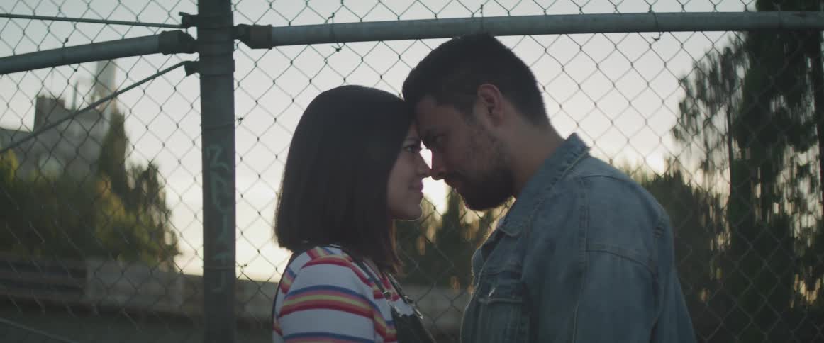 A man and woman gaze into each others eyes next to a chain link fence
