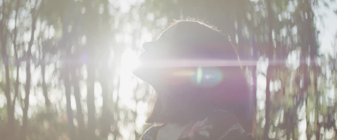 Woman breathes and looks around while the sun streams through the trees in the forest