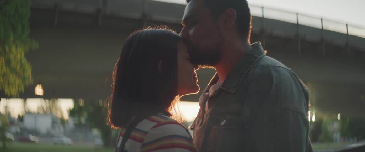 A bearded man kisses a woman on the forehead as the sun sets