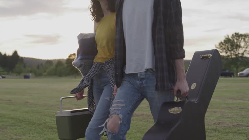 A man with a guitar case and a woman with a cooler walking in a grassy field holding hands