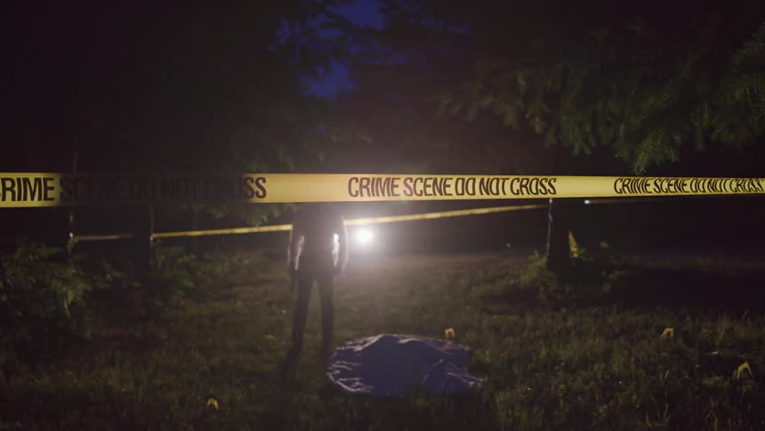 A police officer holding a gun walks past a tarp on the ground in an area marked with crime scene tape