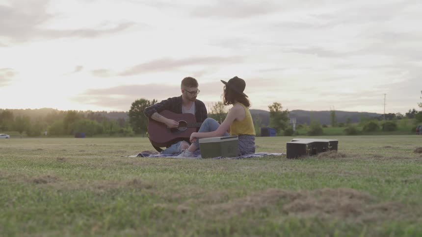 A man plays an acoustic guitar while sitting on a grassy lawn with a woman