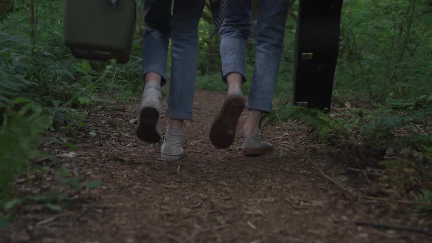 Two females walk along a forest path carrying a cooler and a guitar case