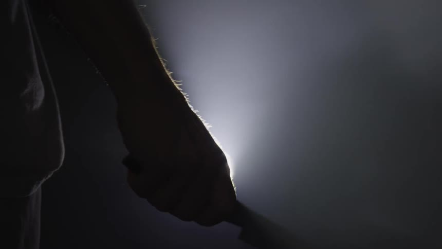 A man standing in the darkness before a flood light while twirling a knife with one day