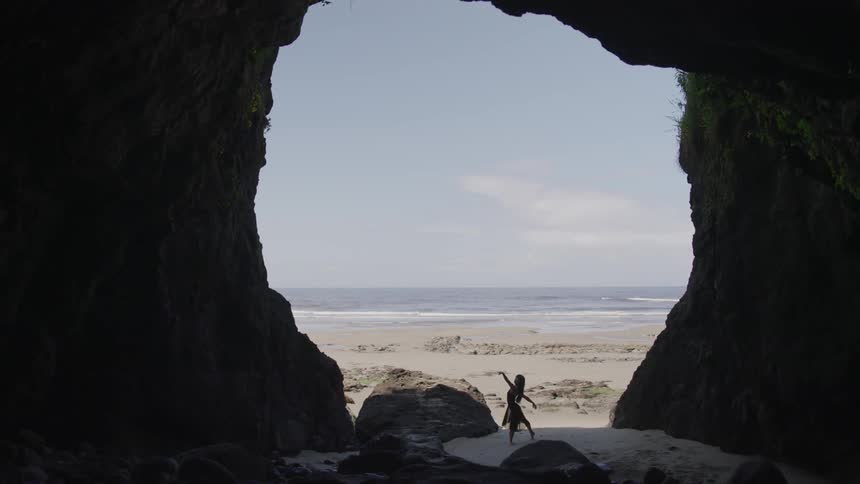 Woman performs dance moves as she stands in the opening of a cave near the ocean