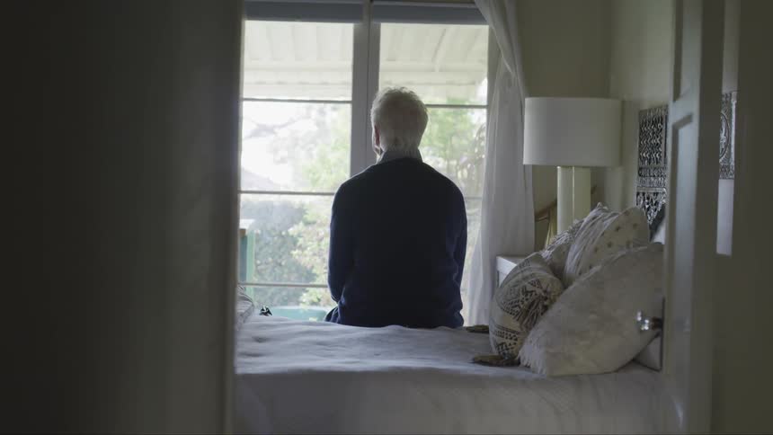 Older person sits on side of bed looking out the window