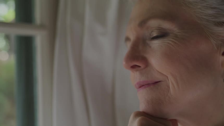 An elderly woman grinning while looking out of a window contemplatively