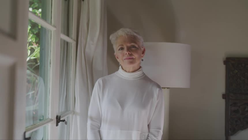 Old woman wearing white pullover looks out the window and turns to the camera in the bedroom