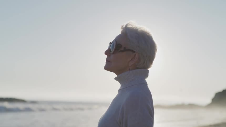 An elderly woman stands on the beach and looks towards the water