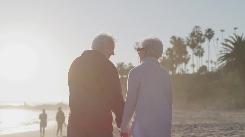 An elderly couple walk across a beach while holding hands