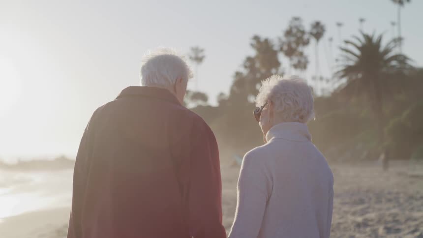 An elderly man and woman walk along the beach holding hands