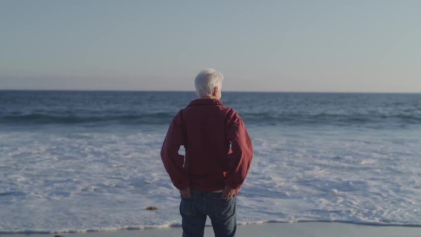 An elderly man stands on a beach with his hands in his back pockets