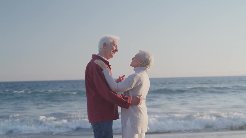 An elderly couple holds each other close as they slowly dance on the beach in front of crashing waves