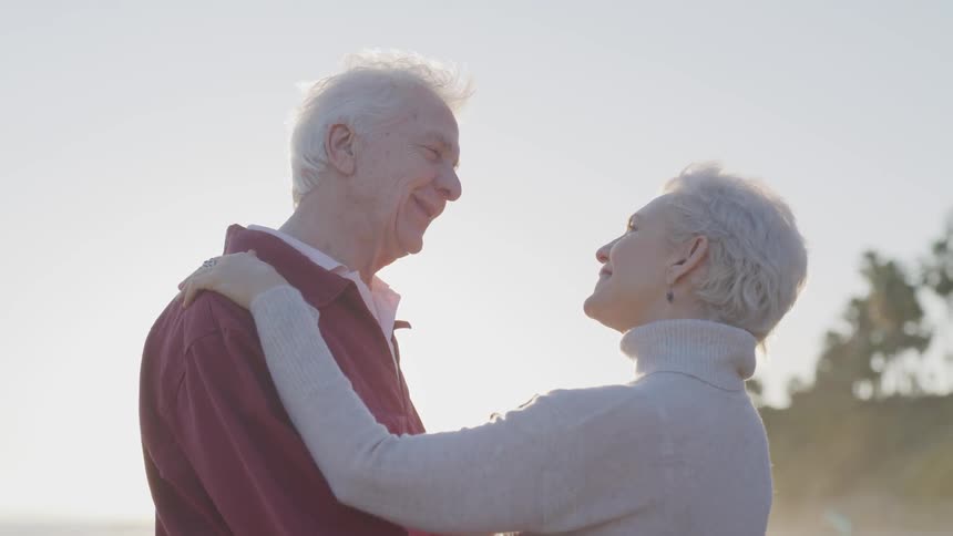 Old man and woman hold and look at each other on the beach at sunset next to the sea
