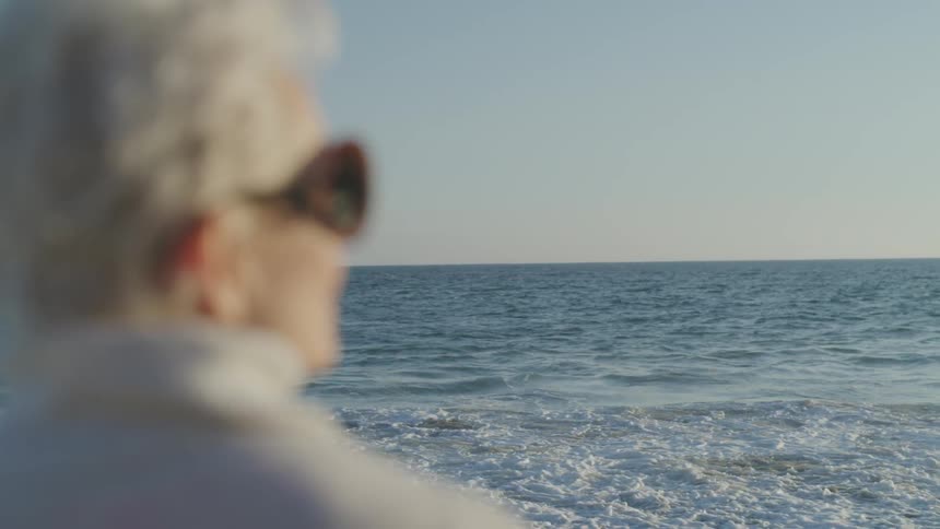 Old woman looks at sea on the sand beach at sunset