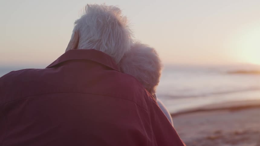 Old man and woman cuddle each other on the beach at sunset next to the sea