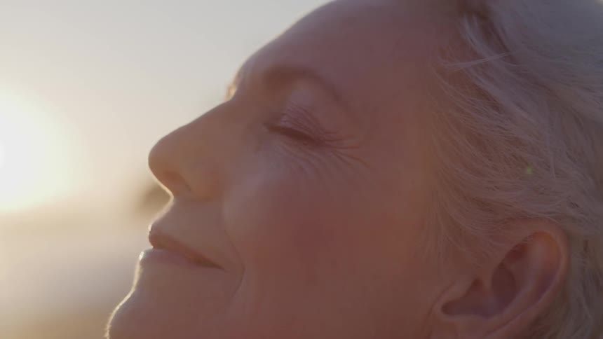 An elderly woman looks up towards the sun with her eyes closed