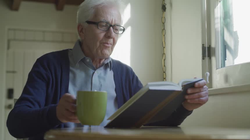 An elderly man is drinking coffee from a green mug while he reads a book