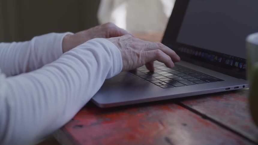 An elderly woman types on a laptop at an old table
