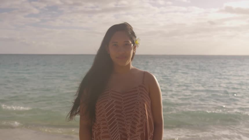 A native woman with a flower in her hair smiles while she stands on a beach