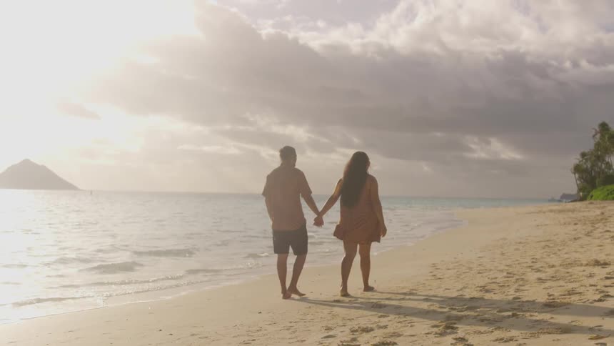 A man and woman hold hands while walking barefoot on a sandy beach
